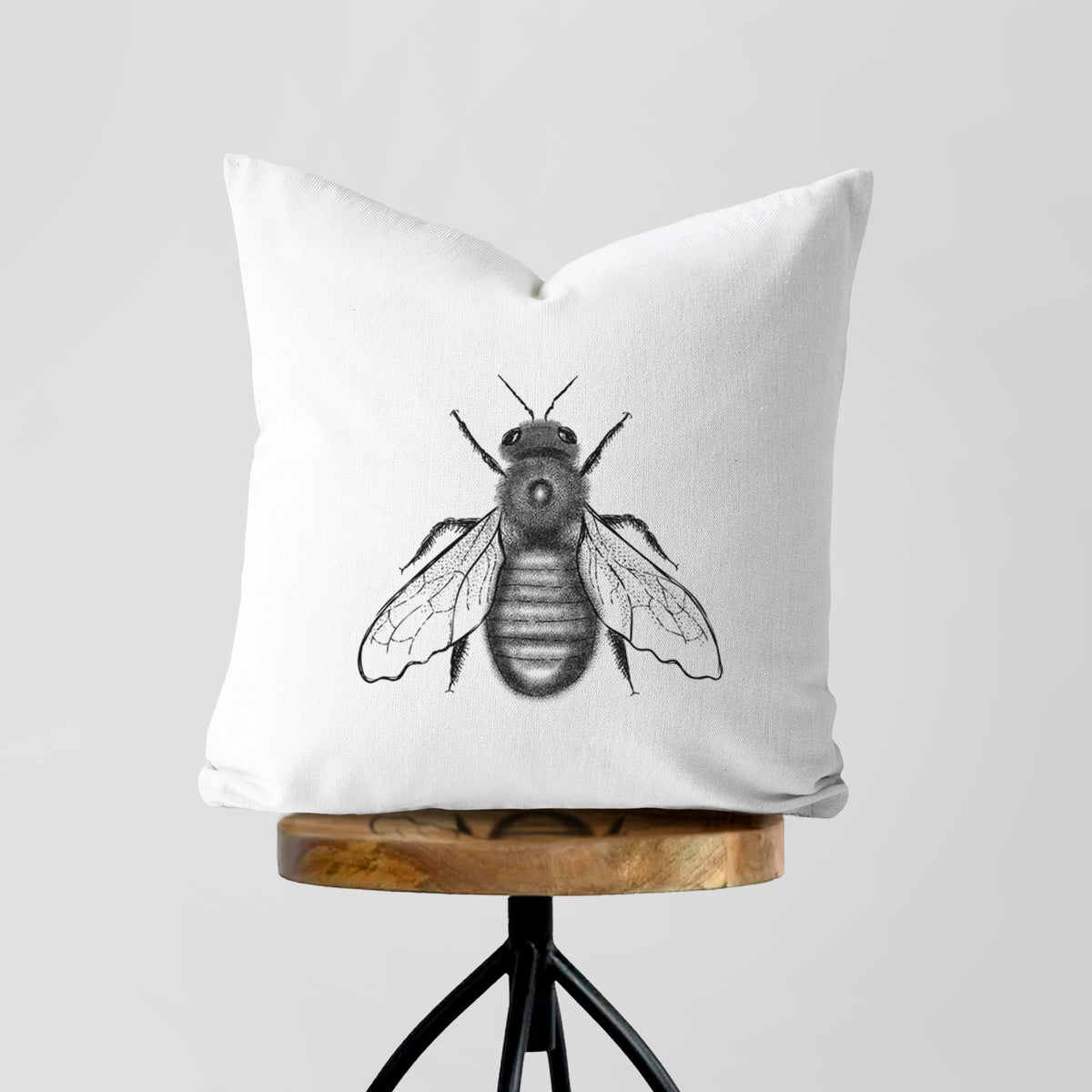 White pillow cover printed with a detailed black ink illustration of an eastern carpenter bee with its wings spread, seen from above, on a wooden stool.