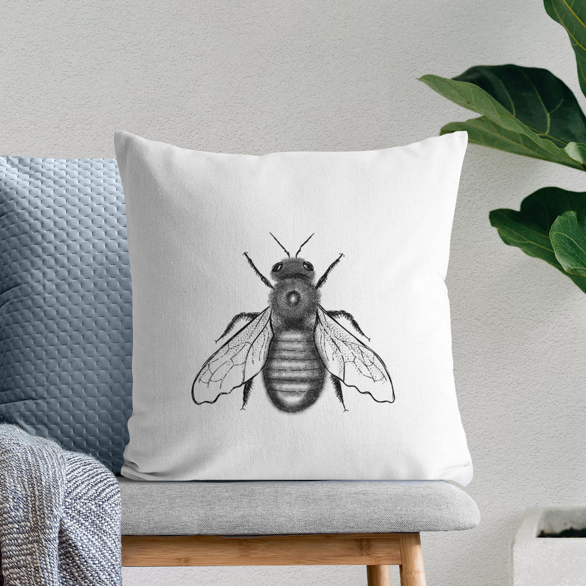 White pillow cover printed with a detailed black ink illustration of an eastern carpenter bee with its wings spread, seen from above, on a bench beside plants.