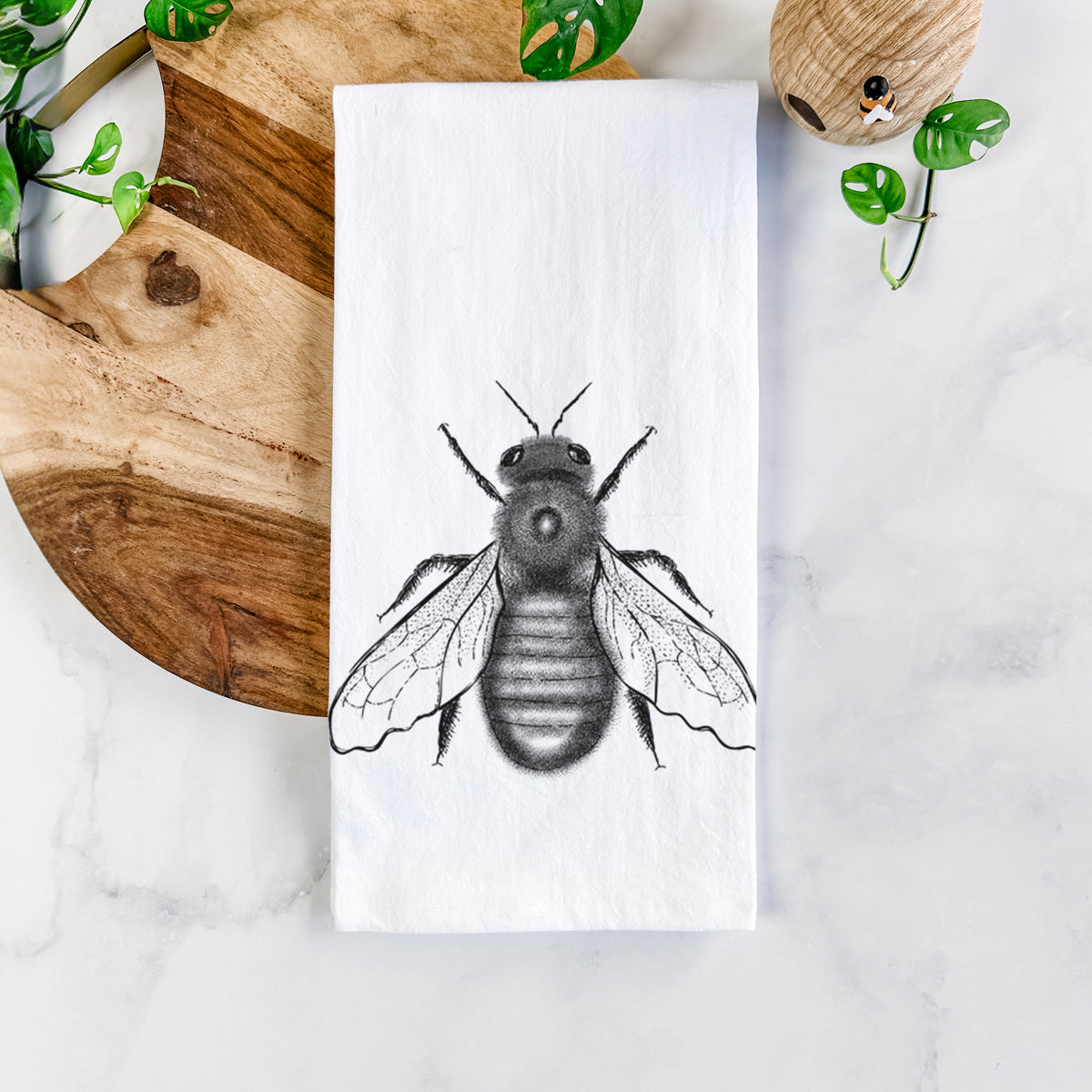 White cotton tea towel printed with a detailed black ink illustration of an eastern carpenter bee with its wings spread, seen from above, on a marble counter beside a round wooden cutting board and greenery.