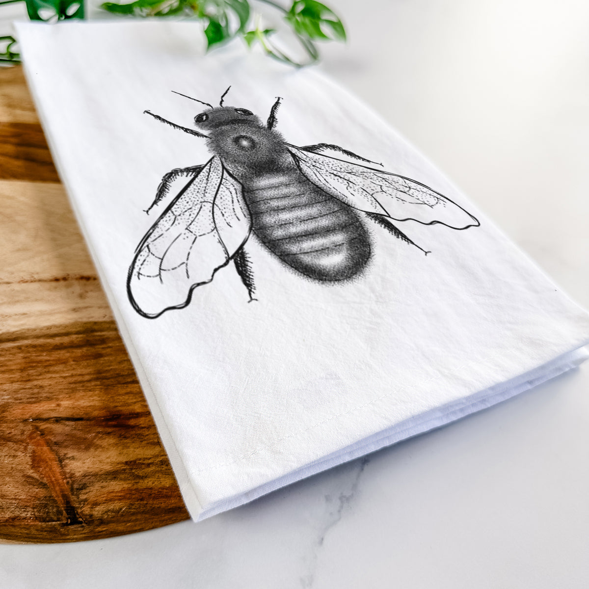 Close-up of a folded white tea towel on a wooden board, showing a detailed black ink illustration of an eastern carpenter bee with its wings spread, seen from above.
