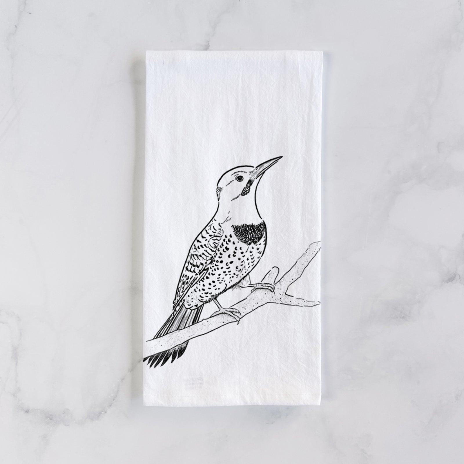 White cotton tea towel printed with a detailed black ink illustration of a yellow-shafted northern flicker perched on a branch, on a marble counter beside a round wooden cutting board and greenery.