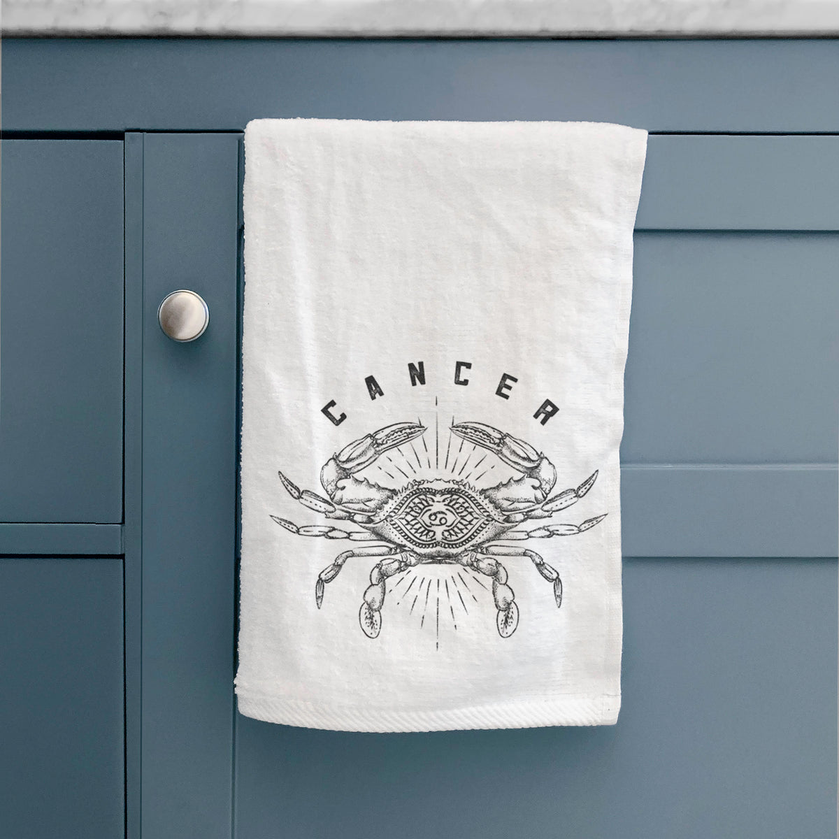 White hand towel printed with a black ink crab with the word Cancer, for the Cancer zodiac sign, hanging on a cabinet door.