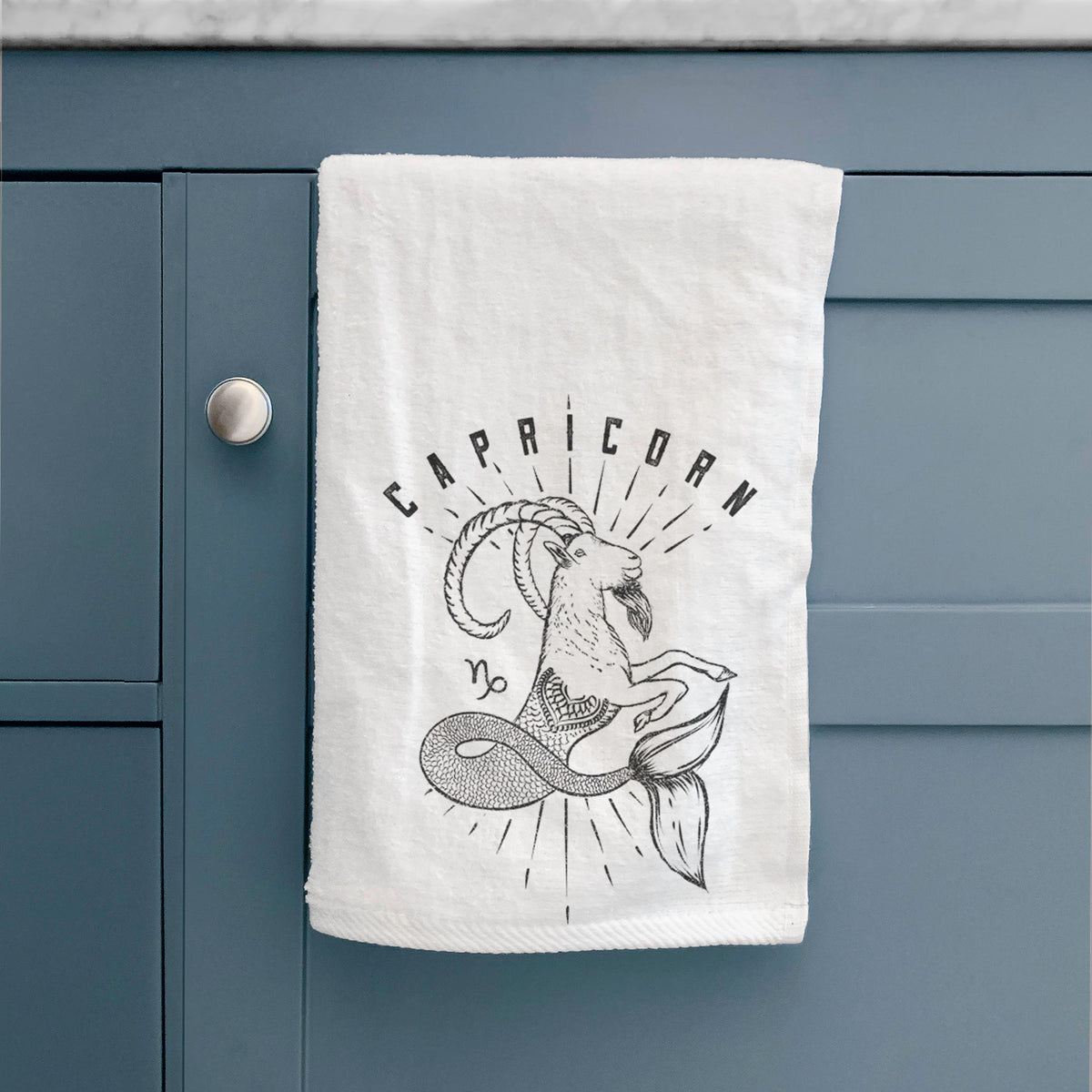 White hand towel printed with a black ink sea-goat with the word Capricorn, for the Capricorn zodiac sign, hanging on a cabinet door.