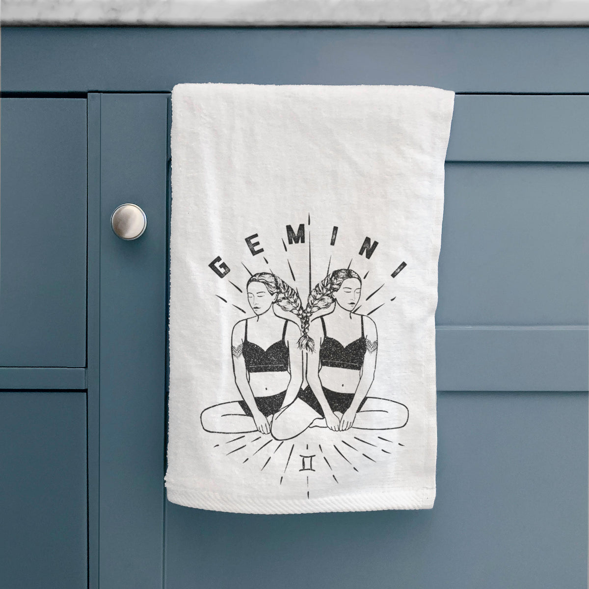 White hand towel printed with black ink twin women sitting back to back with the word Gemini, for the Gemini zodiac sign, hanging on a cabinet door.
