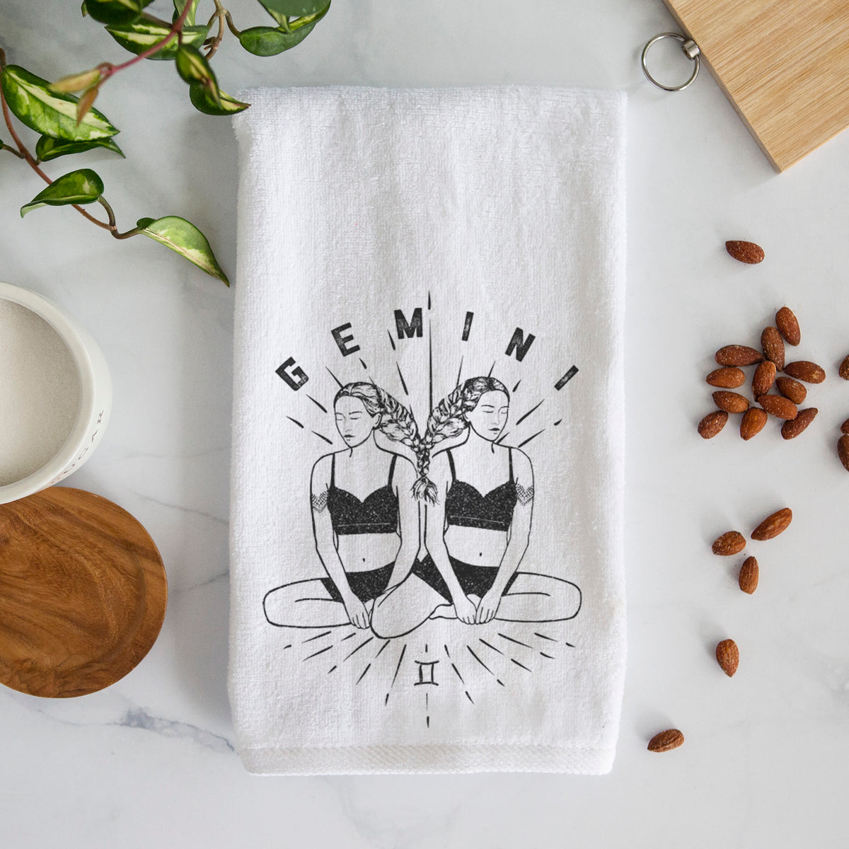 White hand towel printed with black ink twin women sitting back to back with the word Gemini, for the Gemini zodiac sign.