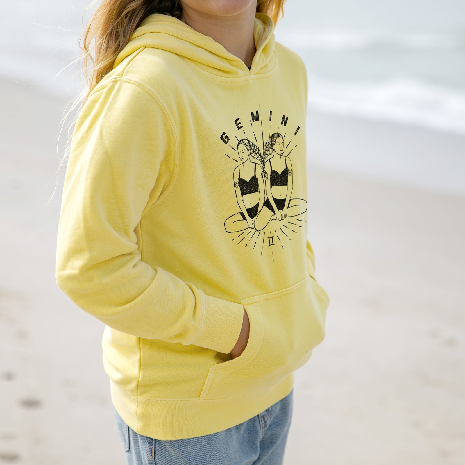 Boy wearing a youth pigment dyed hoodie printed with black ink twin women sitting back to back with the word Gemini, for the Gemini zodiac sign.