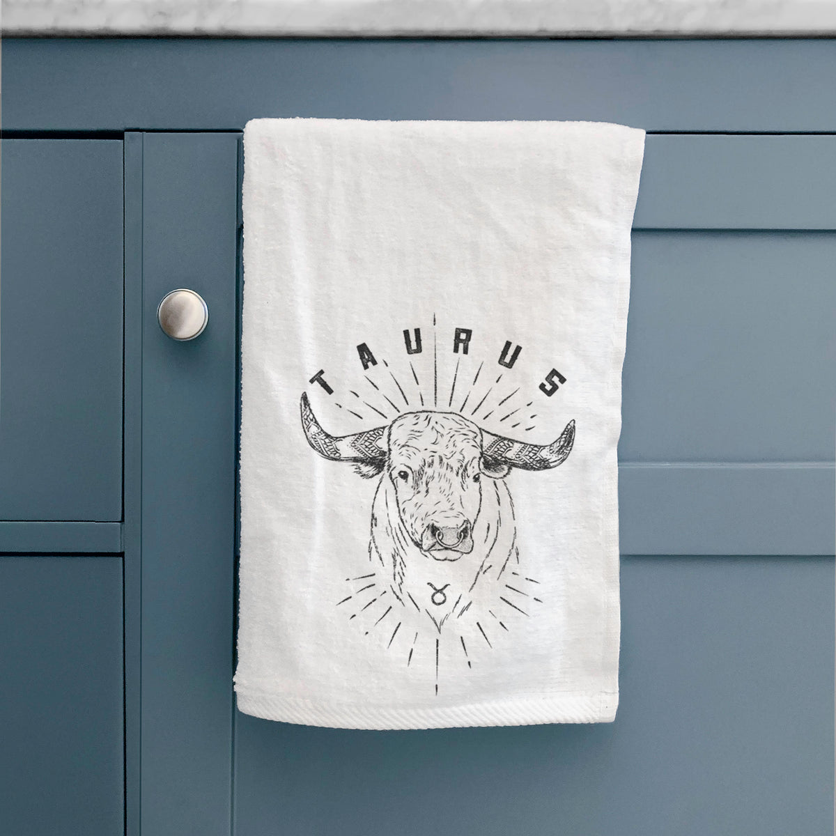 White hand towel printed with a black ink bull's head with the word Taurus, for the Taurus zodiac sign, hanging on a cabinet door.