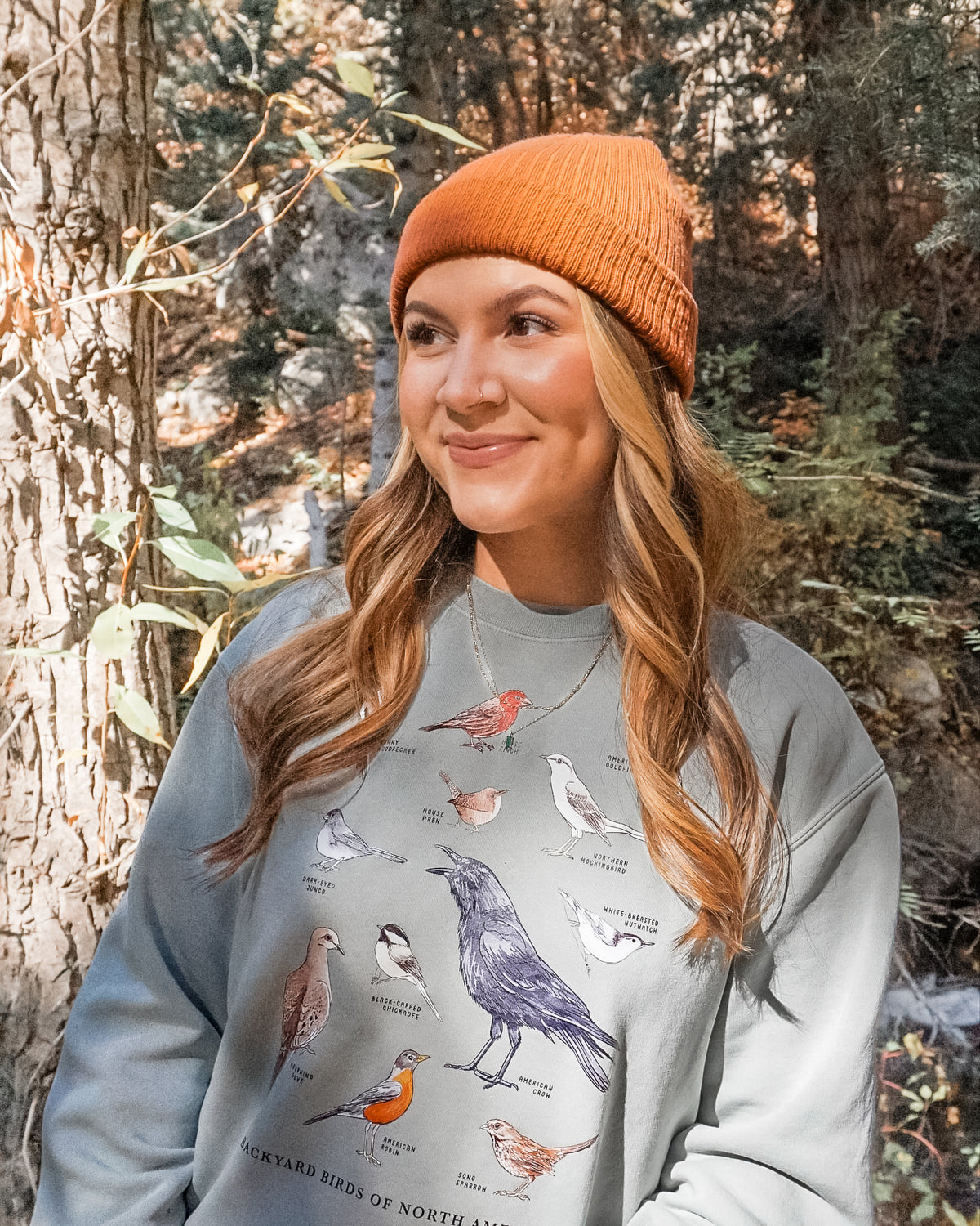 Woman wearing a gray sweatshirt with bird illustrations in a forest setting