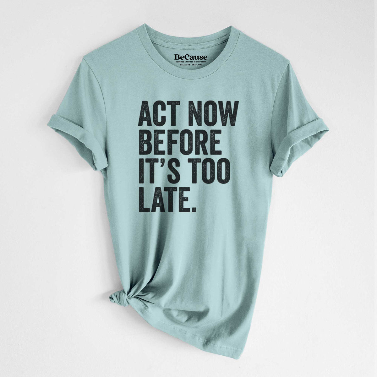 Dusty blue lightweight cotton t-shirt laid flat with the sleeves rolled, printed with bold black words reading Act Now Before It's Too Late.