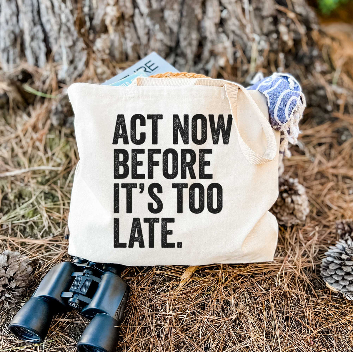 Canvas tote bag printed with bold black words reading Act Now Before It's Too Late, resting on dry grass beside a pair of binoculars.