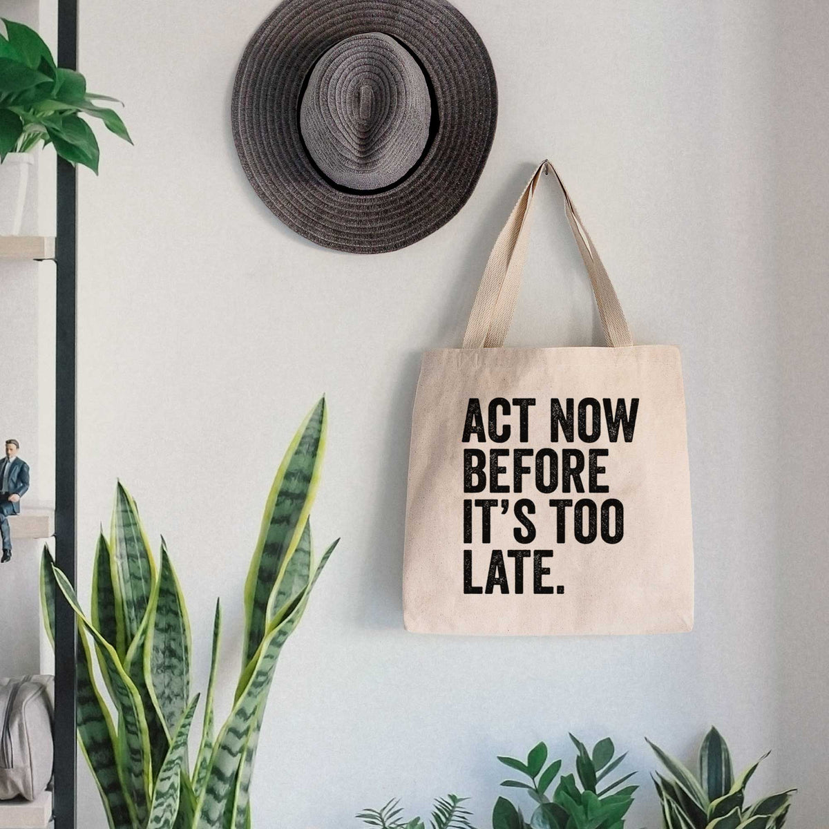 Canvas tote bag printed with bold black words reading Act Now Before It's Too Late, hanging on a wall hook below a hat, next to potted plants.