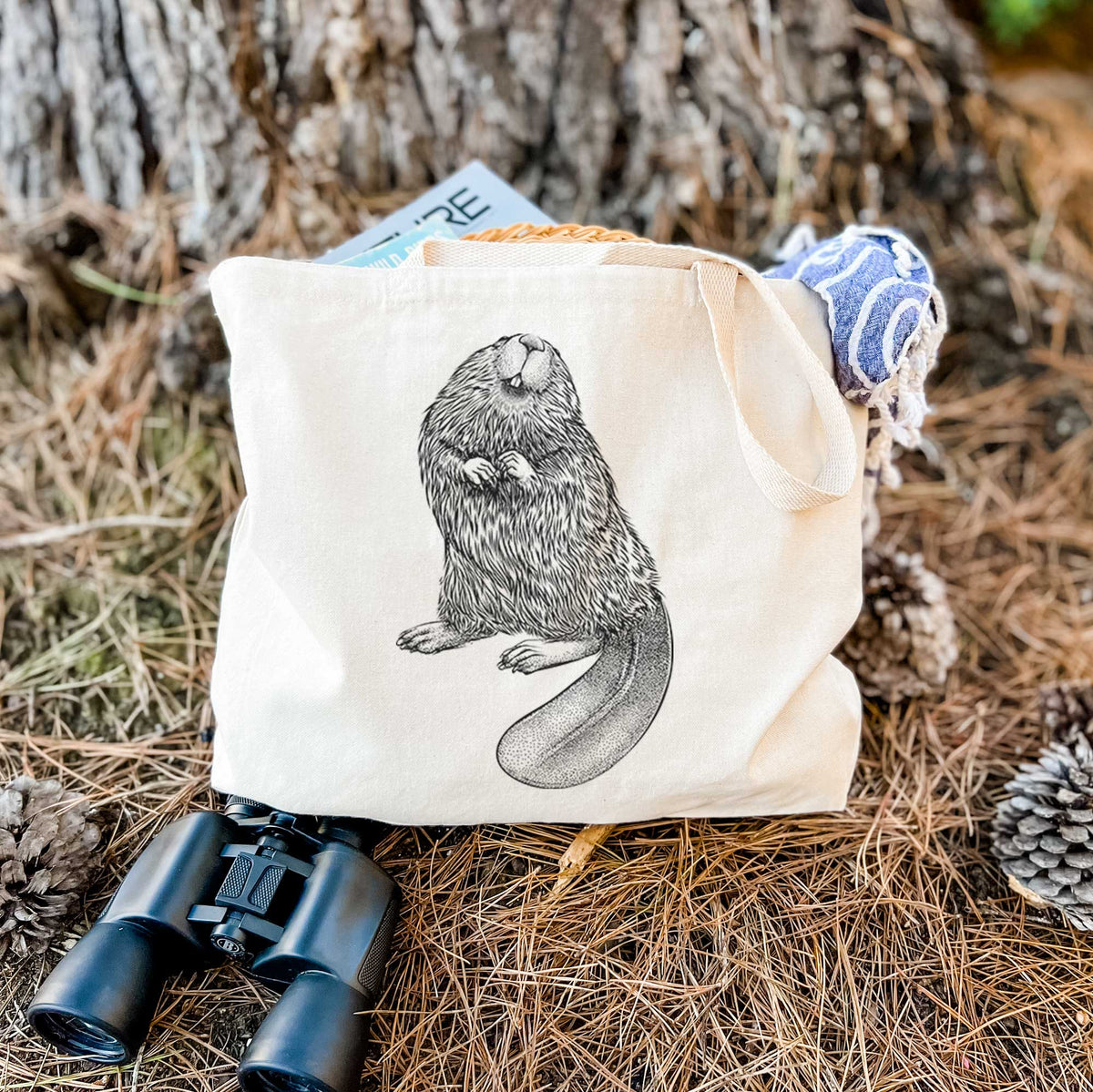 Canvas tote bag printed with a detailed black ink illustration of an American beaver sitting upright with its flat tail, resting on dry grass beside a pair of binoculars.