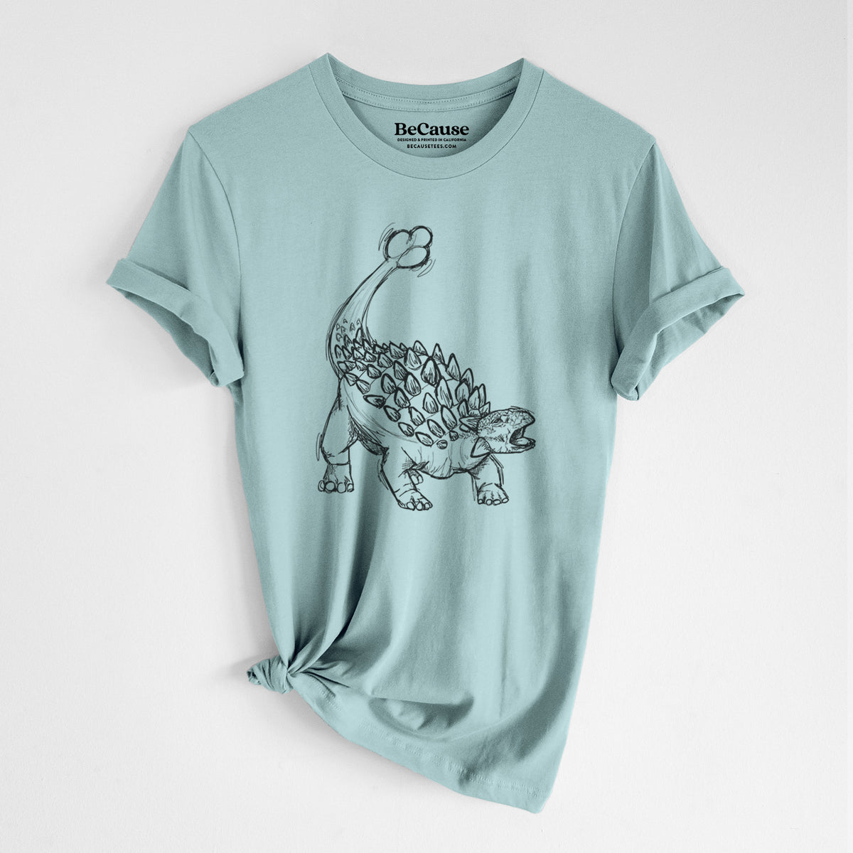 Dusty blue lightweight cotton t-shirt laid flat with the sleeves rolled, printed with a black ink illustration of an Ankylosaurus with its club tail raised.