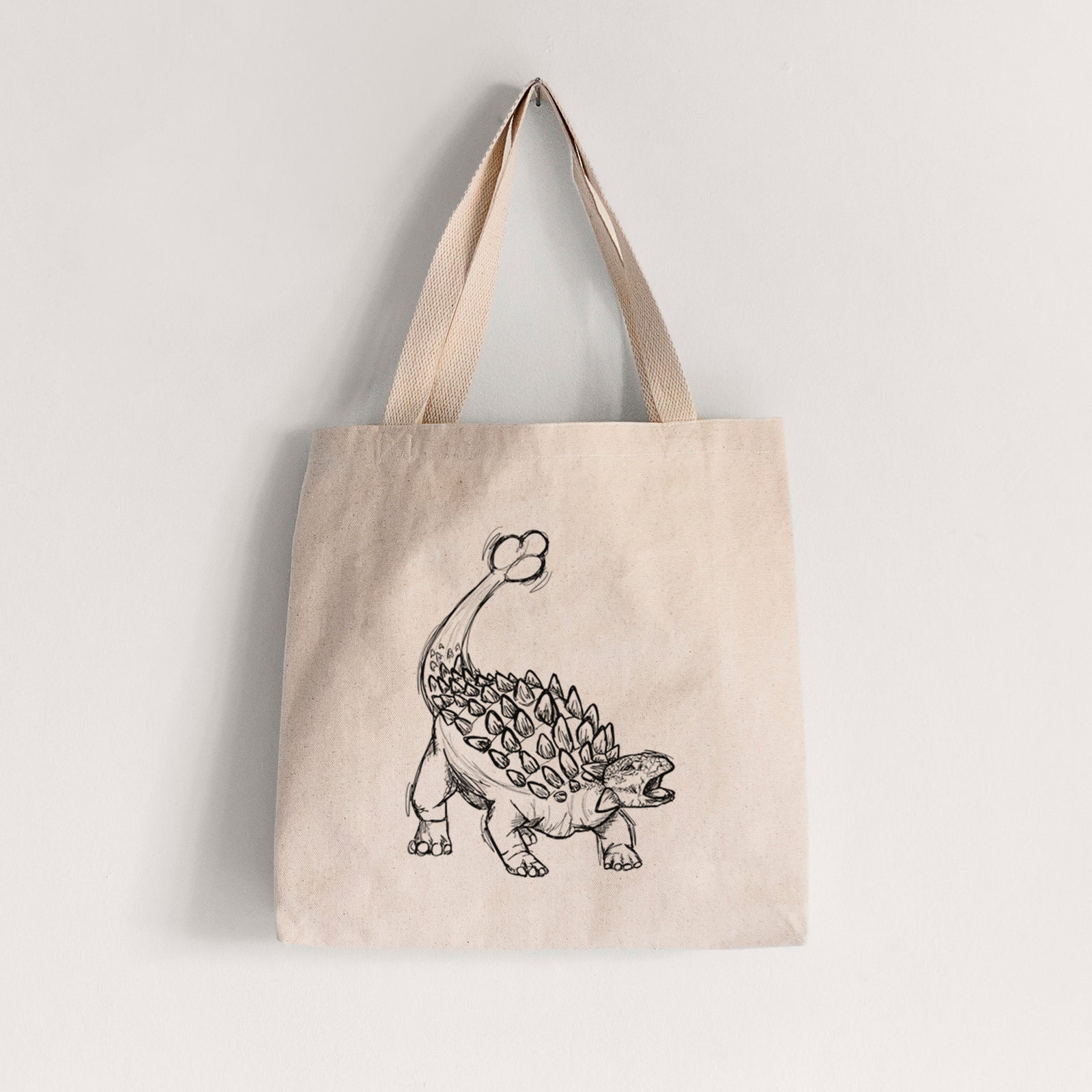 Natural canvas tote bag printed with a black ink illustration of an Ankylosaurus with its club tail raised.