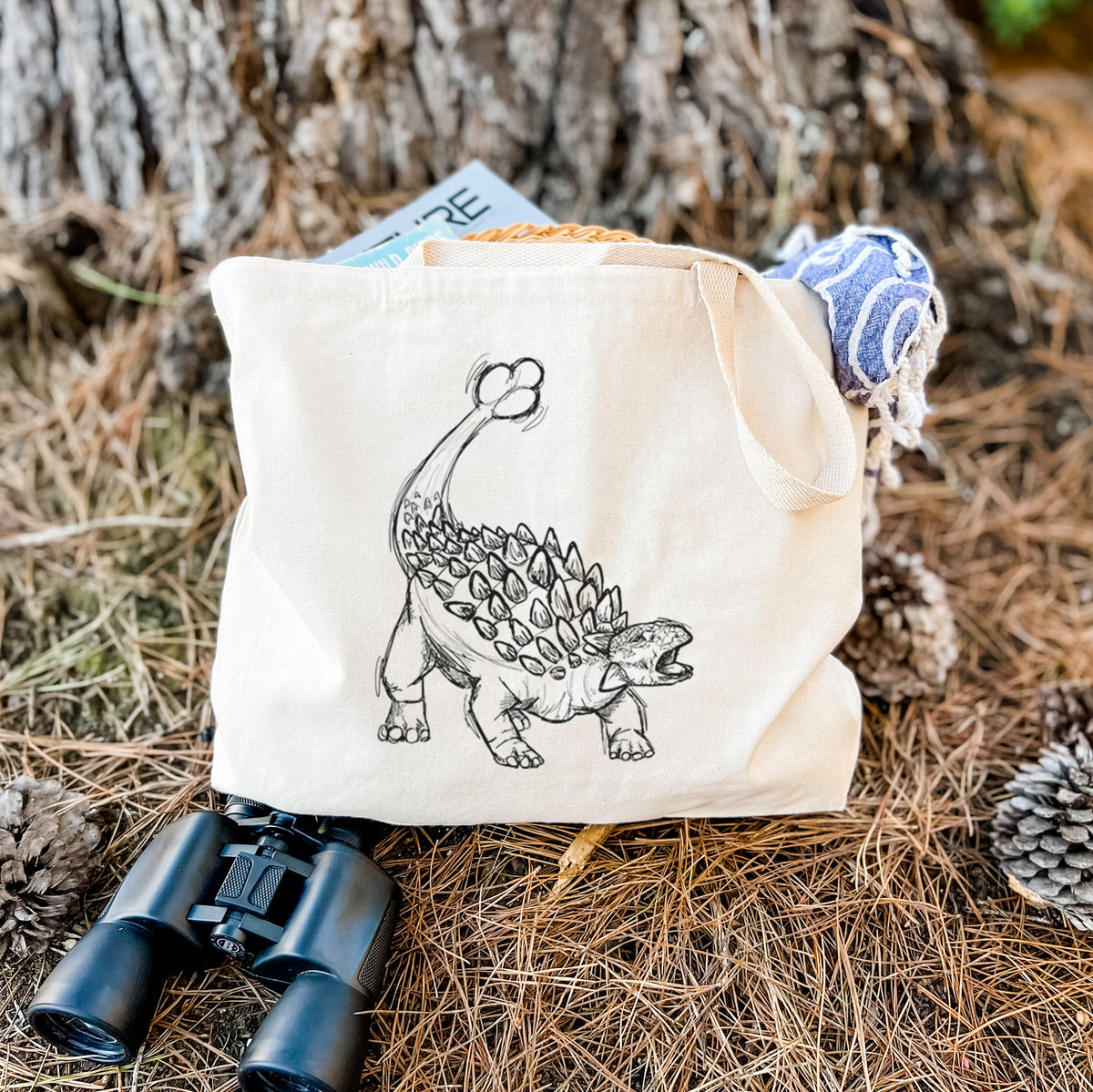 Canvas tote bag printed with a black ink illustration of an Ankylosaurus with its club tail raised, resting on dry grass beside a pair of binoculars.