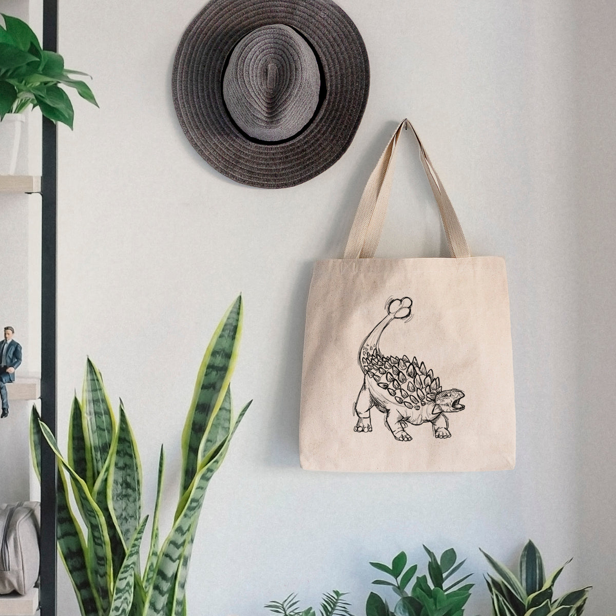 Canvas tote bag printed with a black ink illustration of an Ankylosaurus with its club tail raised, hanging on a wall hook below a hat, next to potted plants.