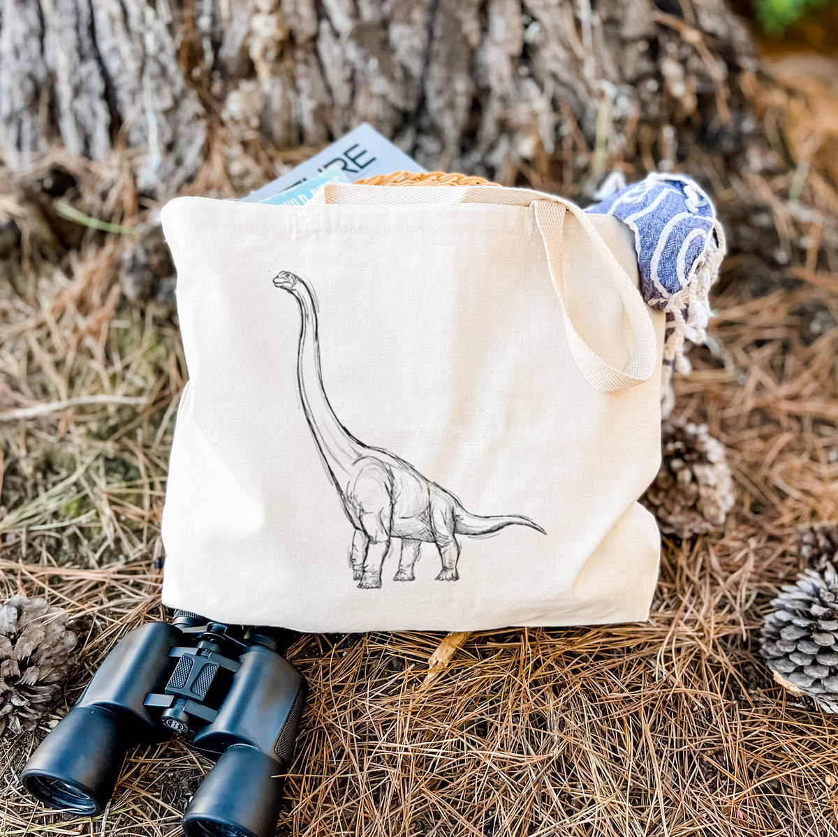 Canvas tote bag printed with a black ink illustration of a long-necked Apatosaurus walking, resting on dry grass beside a pair of binoculars.