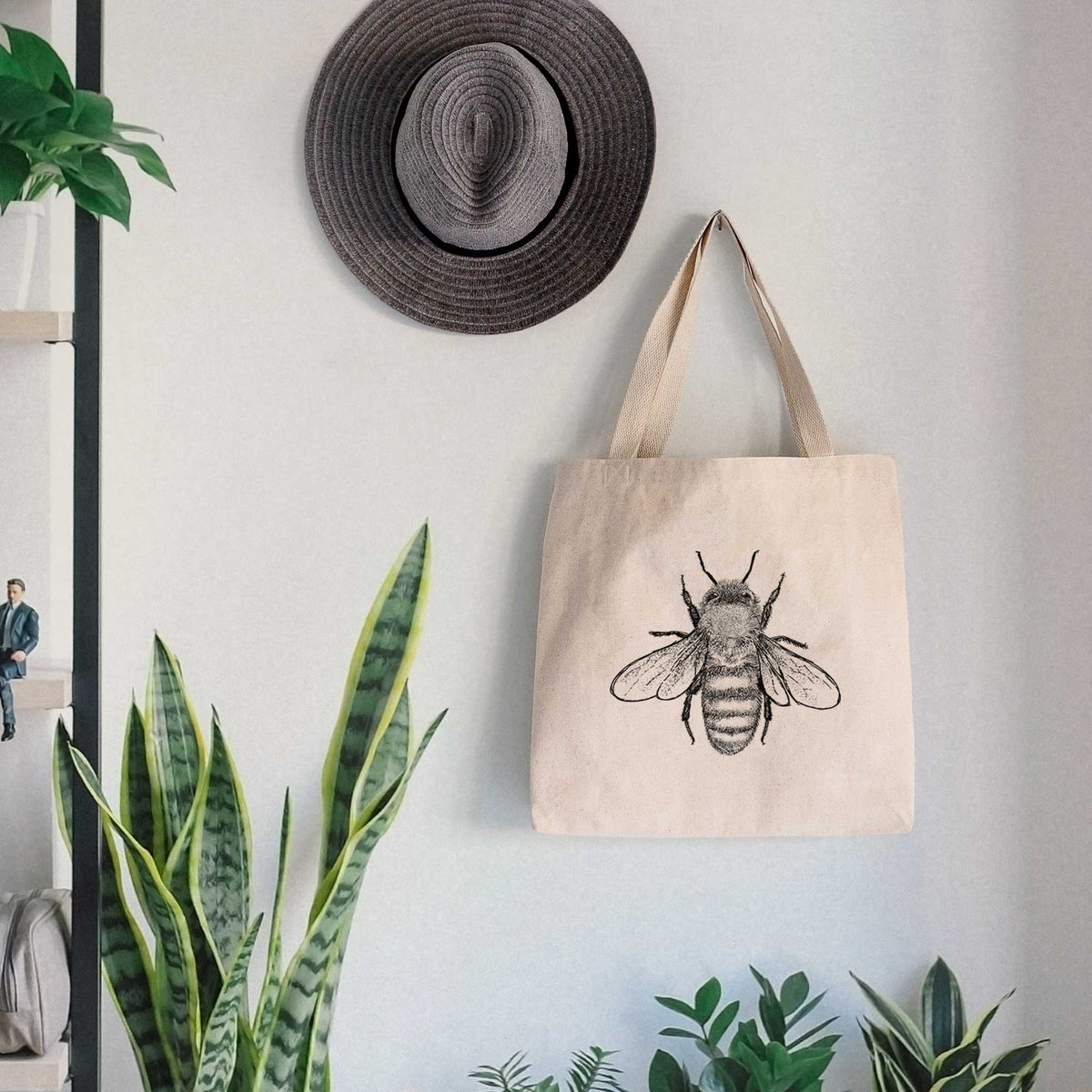 Canvas tote bag printed with a detailed black ink illustration of a honey bee with its wings spread, seen from above, hanging on a wall hook below a hat, next to potted plants.