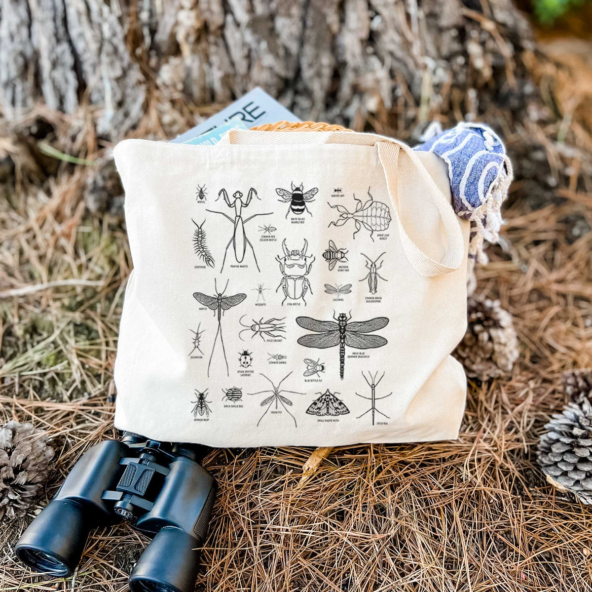 Canvas tote bag printed with a chart of black ink insect and arthropod illustrations, including dragonflies, beetles, spiders and bees, resting on dry grass beside a pair of binoculars.