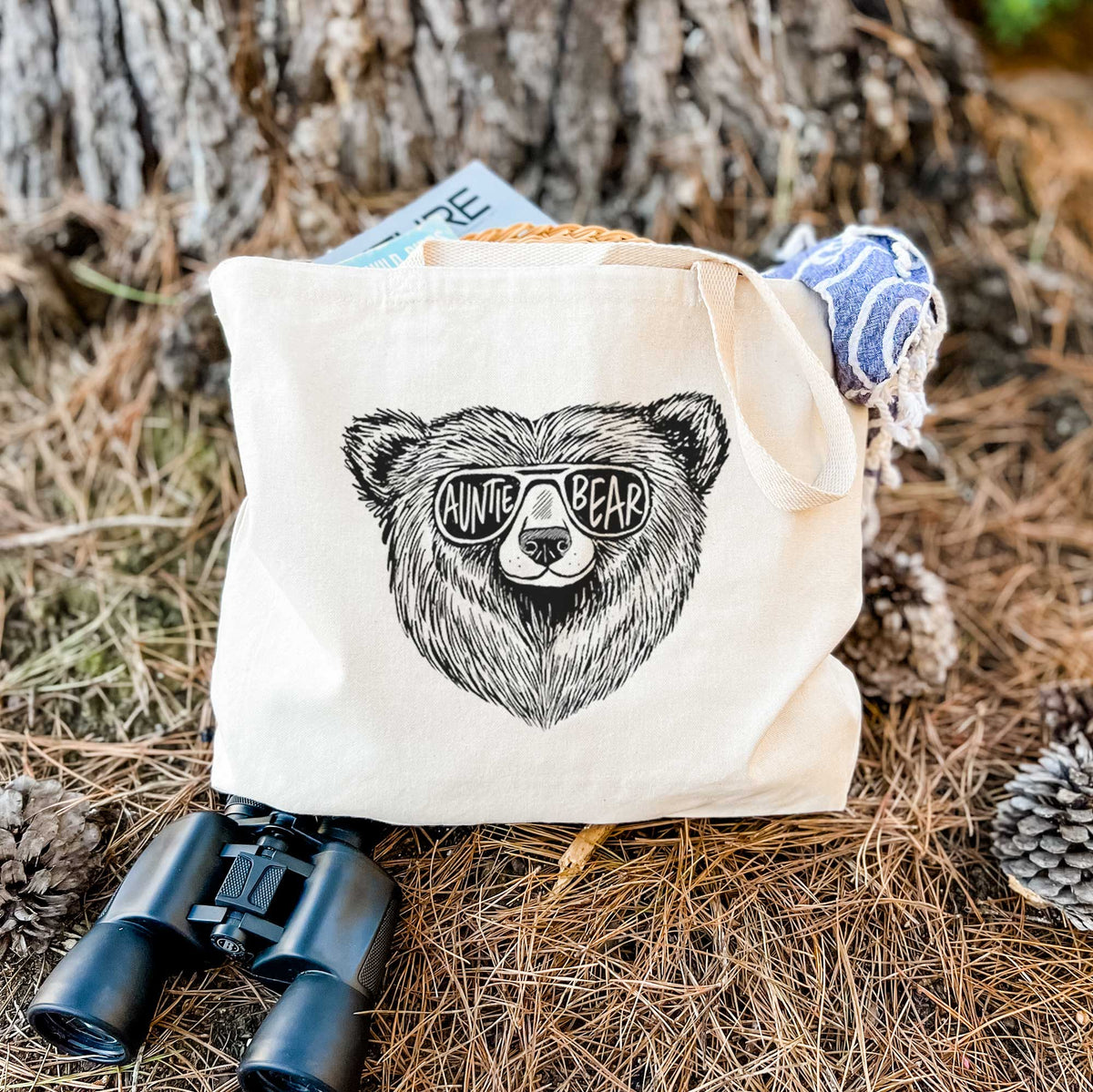 Canvas tote bag printed with a black ink bear's face wearing sunglasses that read Auntie Bear, resting on dry grass beside a pair of binoculars.