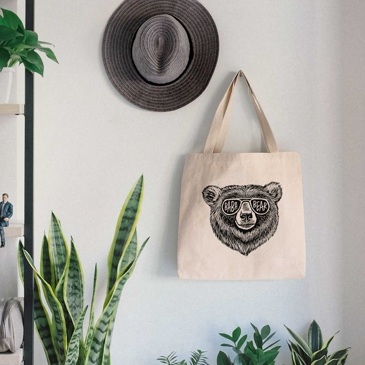 Canvas tote bag printed with a black ink bear's face wearing sunglasses that read Baba Bear, hanging on a wall hook below a hat, next to potted plants.