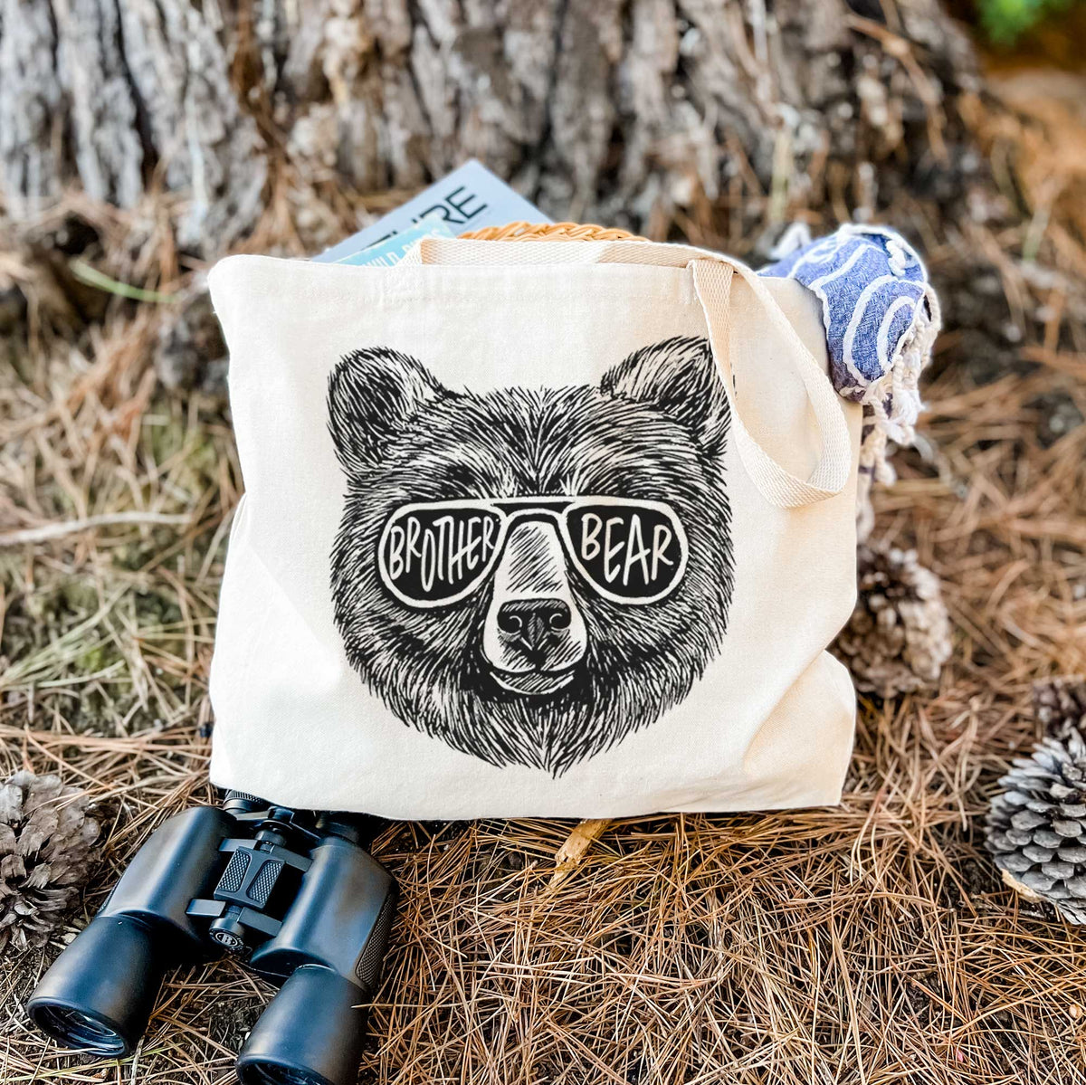 Canvas tote bag printed with a black ink bear's face wearing sunglasses that read Brother Bear, resting on dry grass beside a pair of binoculars.