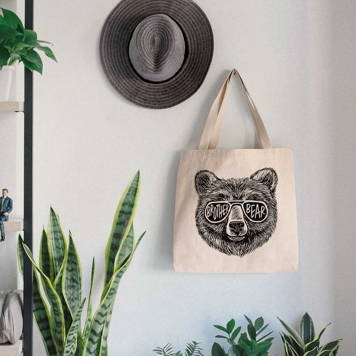 Canvas tote bag printed with a black ink bear's face wearing sunglasses that read Brother Bear, hanging on a wall hook below a hat, next to potted plants.
