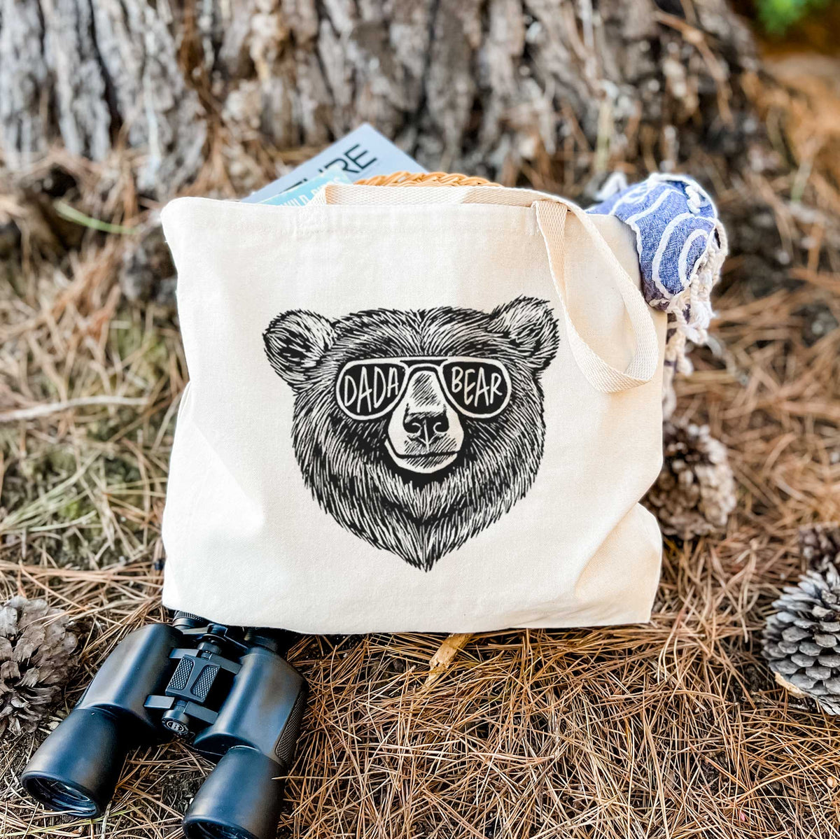 Canvas tote bag printed with a black ink bear's face wearing sunglasses that read Dada Bear, resting on dry grass beside a pair of binoculars.