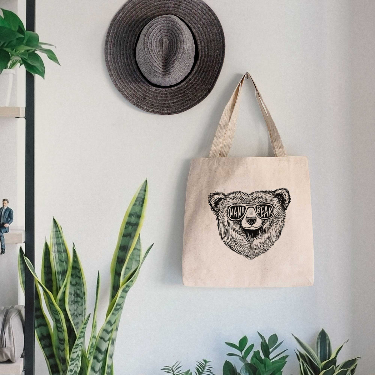 Canvas tote bag printed with a black ink bear's face wearing sunglasses that read Mama Bear, hanging on a wall hook below a hat, next to potted plants.