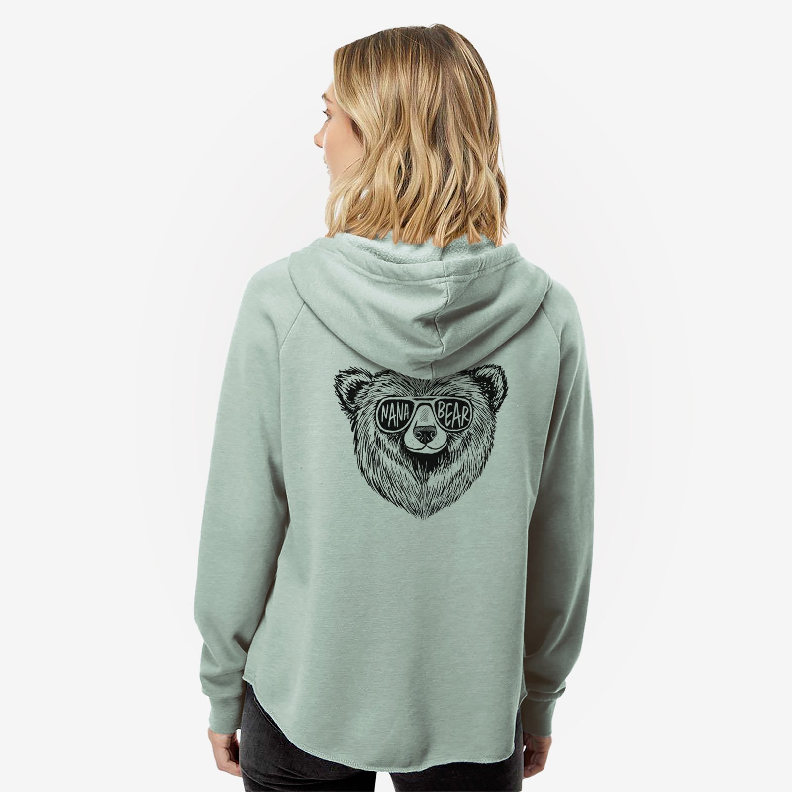 Woman wearing a sage green women's Cali Wave zip-up sweatshirt printed with a black ink bear's face wearing sunglasses that read Nana Bear, seen from behind.