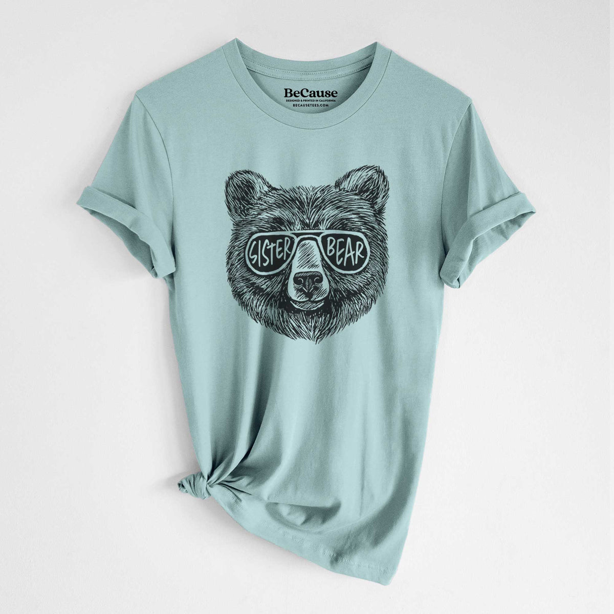 Dusty blue lightweight cotton t-shirt laid flat with the sleeves rolled, printed with a black ink bear's face wearing sunglasses that read Sister Bear.