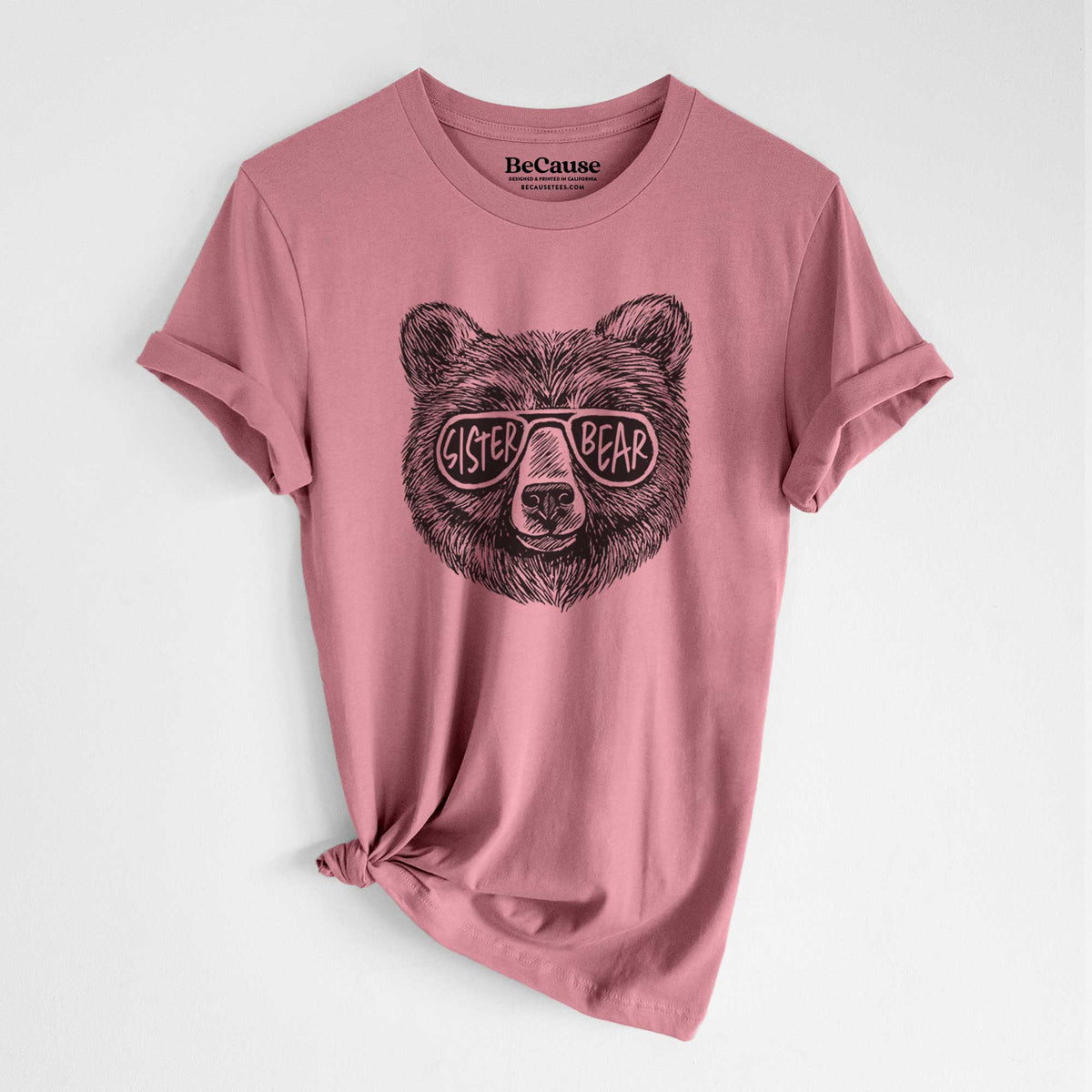 Mauve pink lightweight cotton t-shirt laid flat with the sleeves rolled, printed with a black ink bear's face wearing sunglasses that read Sister Bear.
