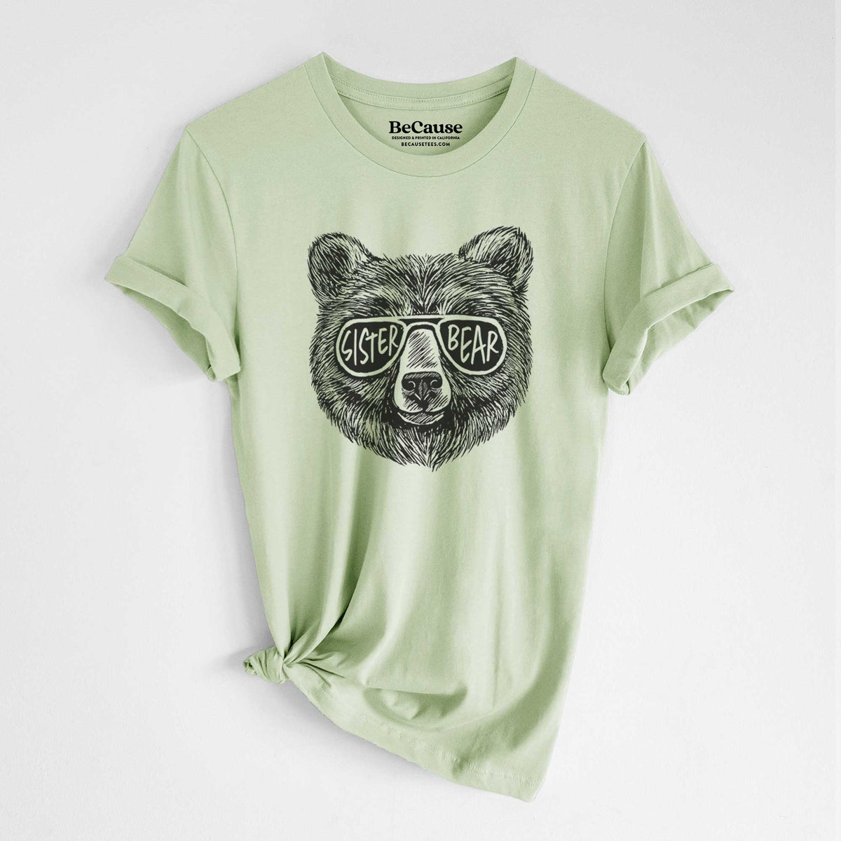 Spring green lightweight cotton t-shirt laid flat with the sleeves rolled, printed with a black ink bear's face wearing sunglasses that read Sister Bear.