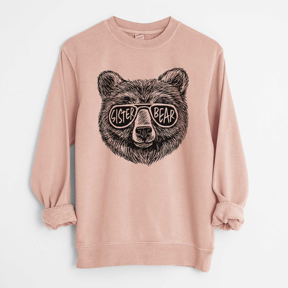 Dusty pink pigment dyed crewneck sweatshirt laid flat with the sleeves rolled, printed with a black ink bear's face wearing sunglasses that read Sister Bear.