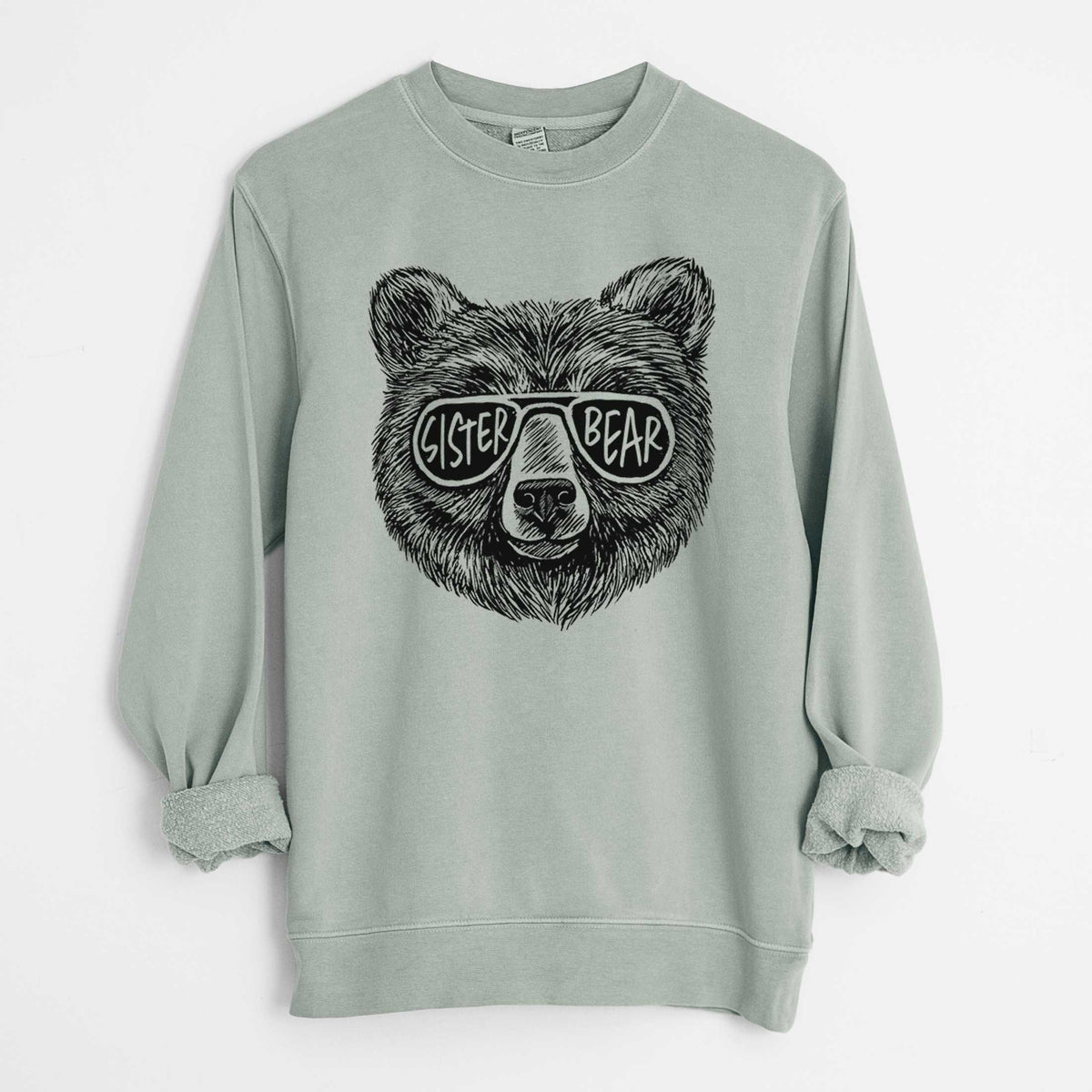 Sage green pigment dyed crewneck sweatshirt laid flat with the sleeves rolled, printed with a black ink bear's face wearing sunglasses that read Sister Bear.