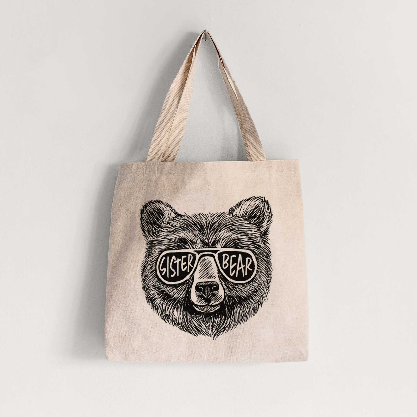 Natural canvas tote bag printed with a black ink bear's face wearing sunglasses that read Sister Bear.