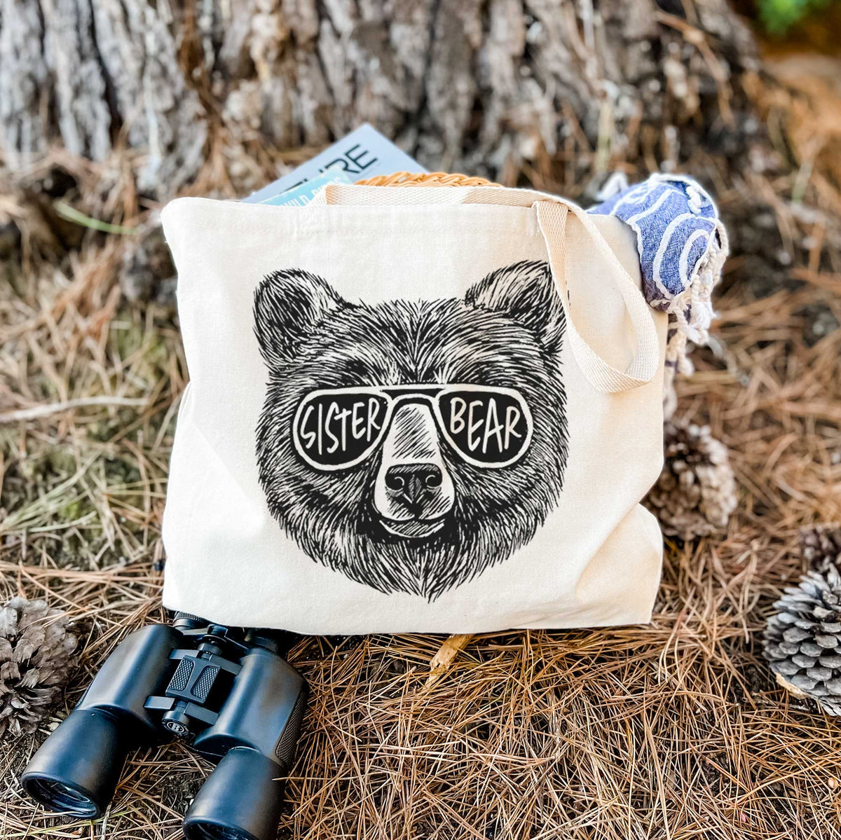 Canvas tote bag printed with a black ink bear's face wearing sunglasses that read Sister Bear, resting on dry grass beside a pair of binoculars.