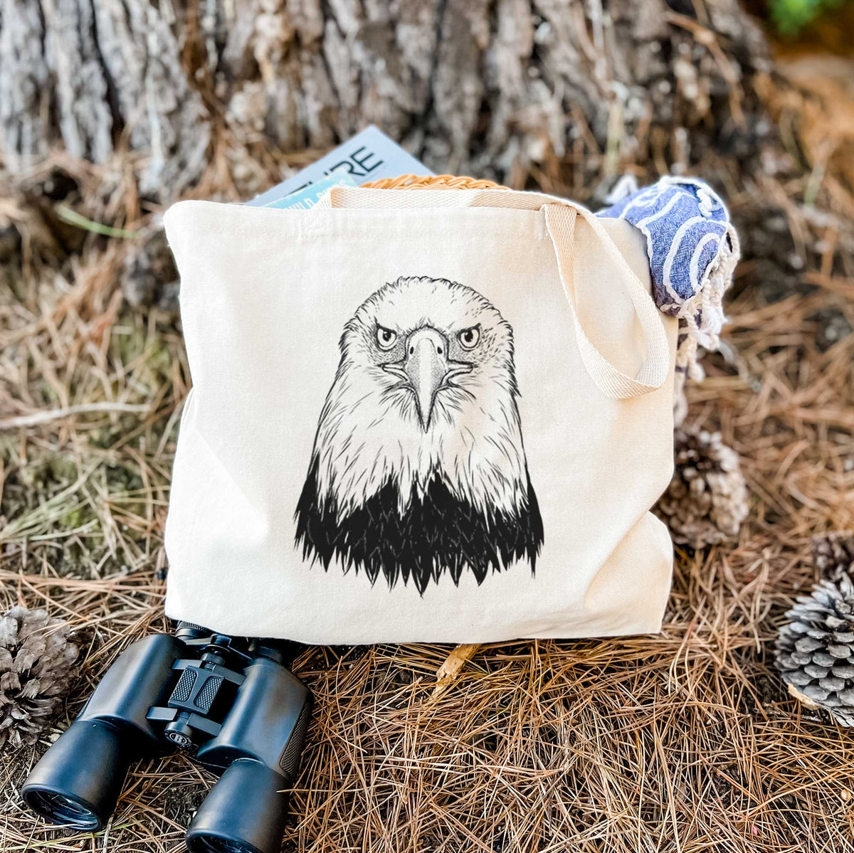 Canvas tote bag printed with a detailed black ink portrait of a bald eagle facing forward, resting on dry grass beside a pair of binoculars.