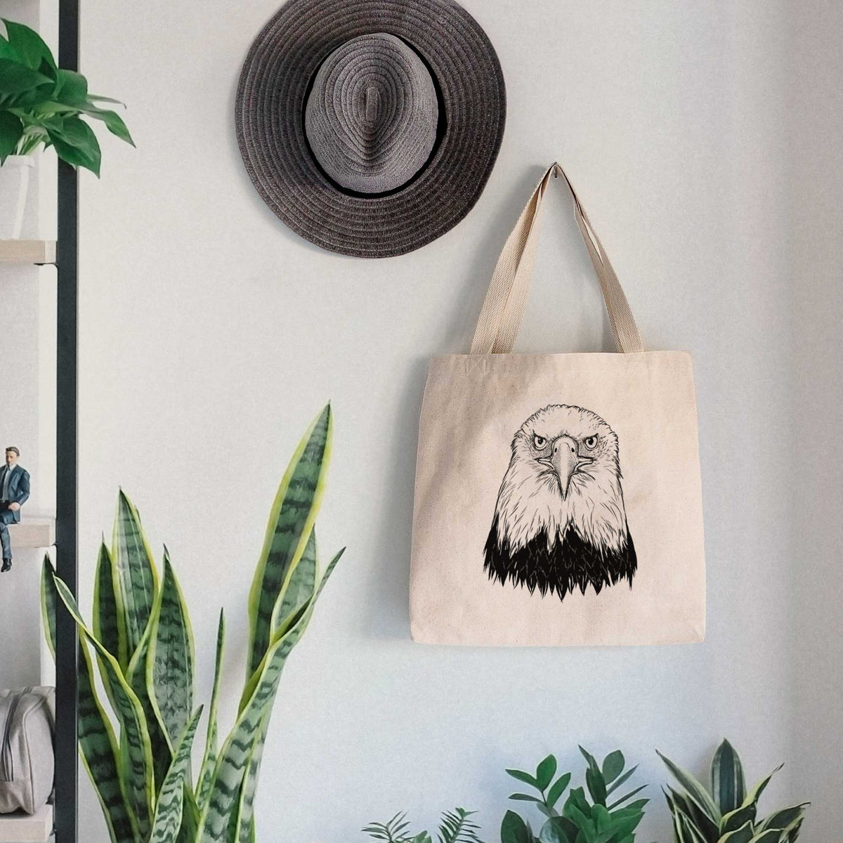 Canvas tote bag printed with a detailed black ink portrait of a bald eagle facing forward, hanging on a wall hook below a hat, next to potted plants.