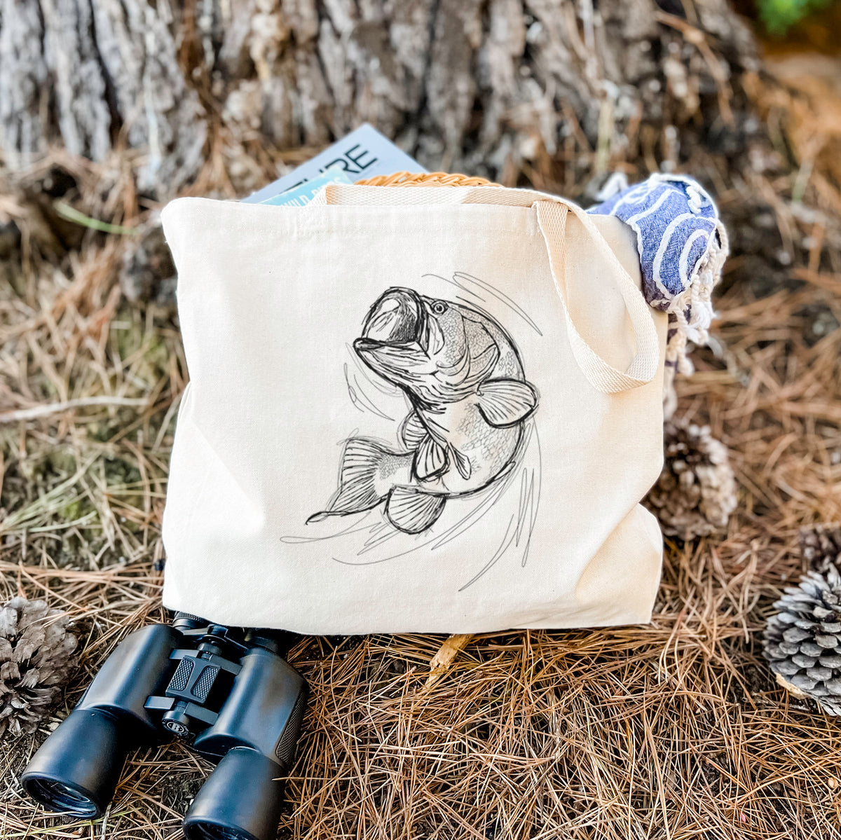 Canvas tote bag printed with a detailed black ink illustration of a largemouth bass leaping with its mouth open, resting on dry grass beside a pair of binoculars.