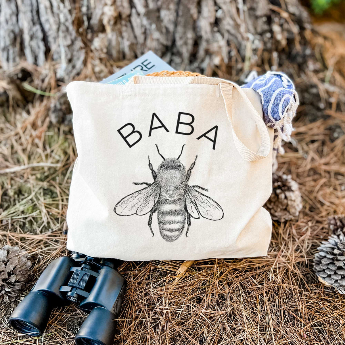 Canvas tote bag printed with a black ink honey bee beneath the word Baba, resting on dry grass beside a pair of binoculars.
