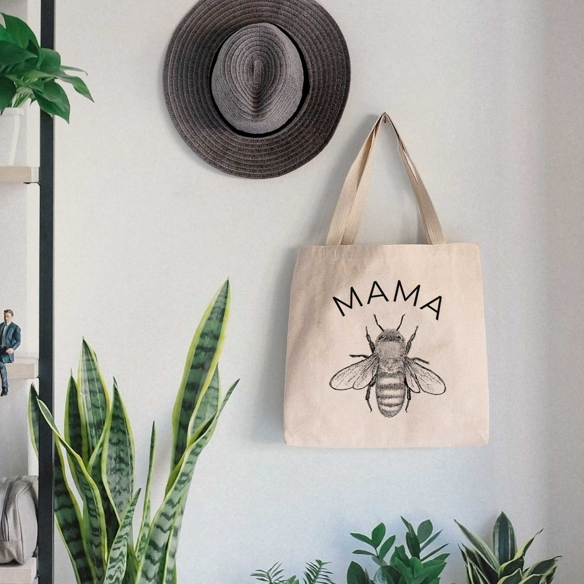 Canvas tote bag printed with a black ink honey bee beneath the word Mama, hanging on a wall hook below a hat, next to potted plants.