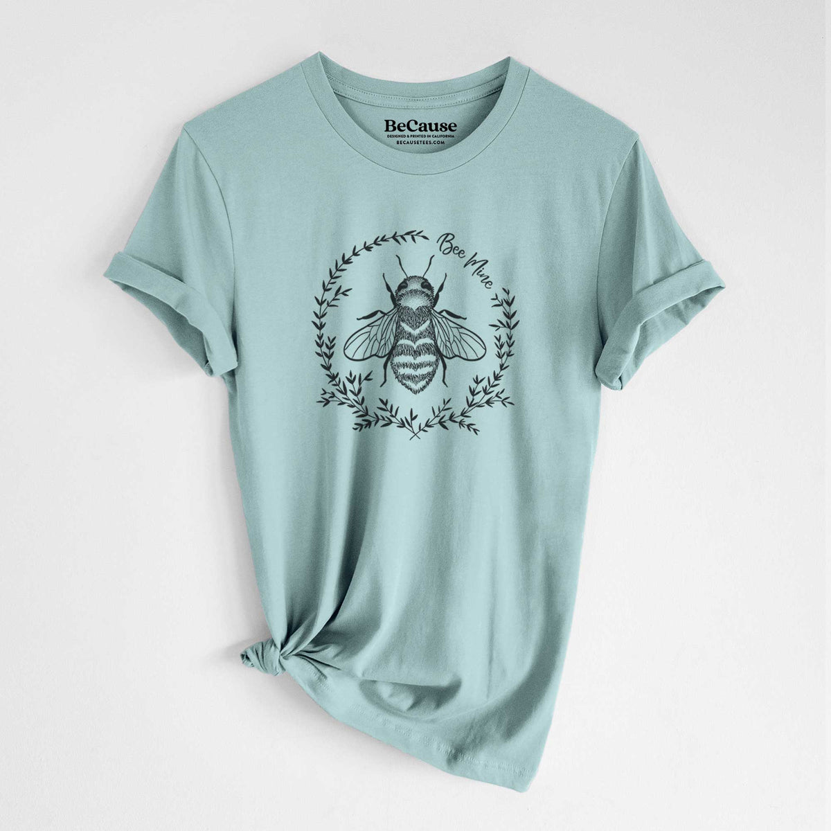 Dusty blue lightweight cotton t-shirt laid flat with the sleeves rolled, printed with a black ink bee inside a wreath of leaves with the words Bee Mine.