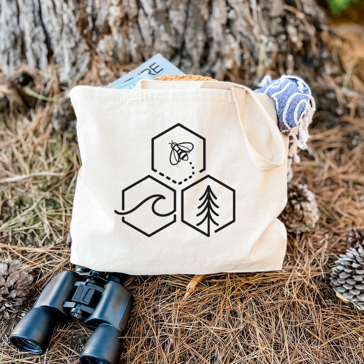 Canvas tote bag printed with a black line art design of three hexagons holding a bee, an ocean wave and a pine tree, resting on dry grass beside a pair of binoculars.