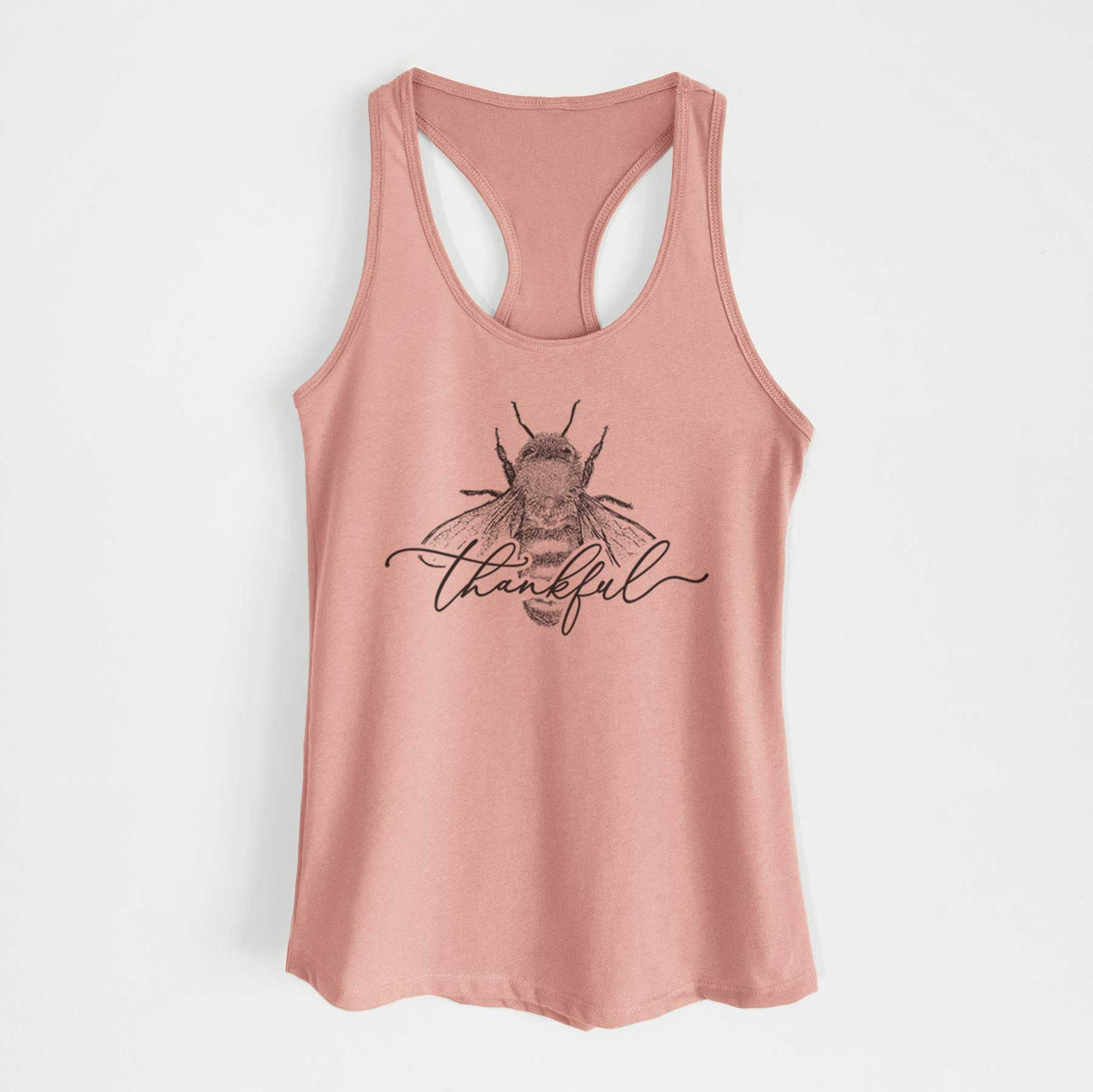 Desert pink women's racerback tank top laid flat, printed with a black ink bee with the word thankful written in script across it.