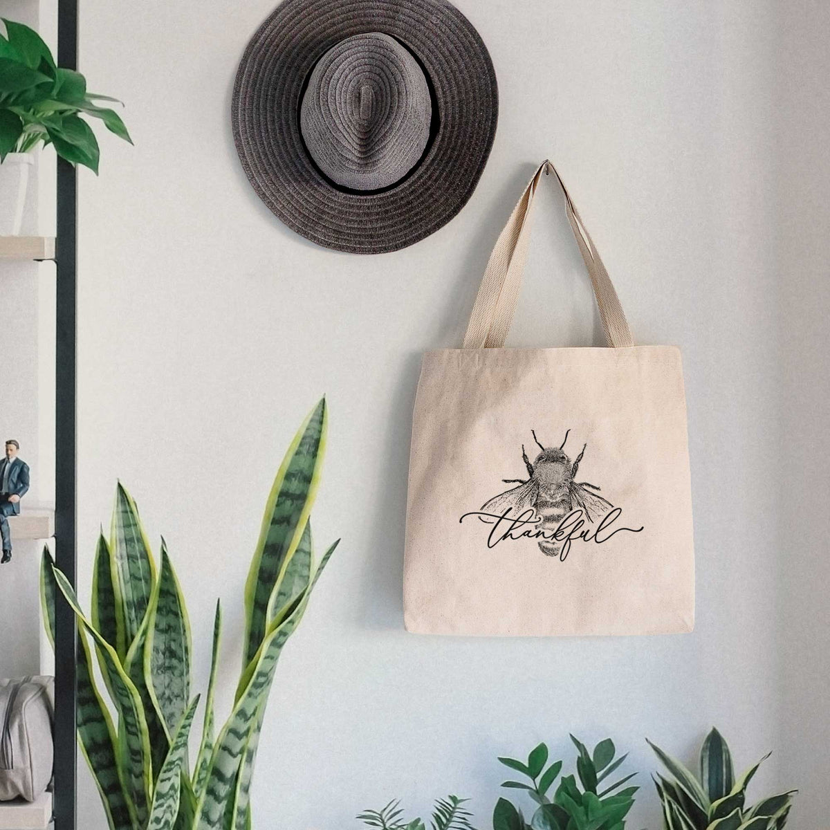 Canvas tote bag printed with a black ink bee with the word thankful written in script across it, hanging on a wall hook below a hat, next to potted plants.