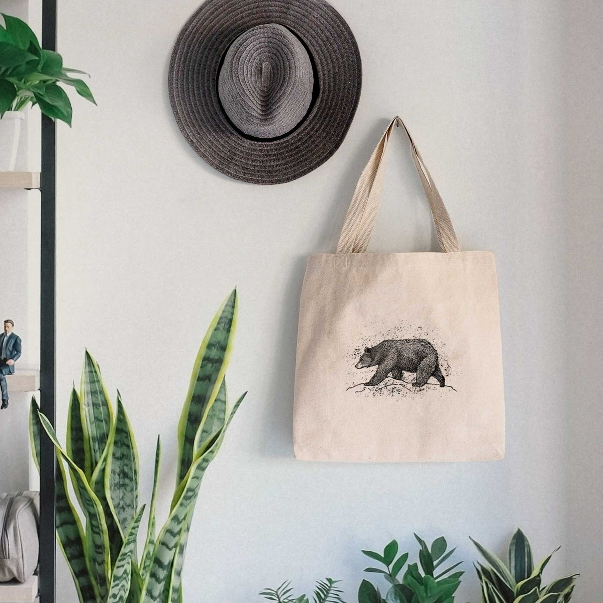 Canvas tote bag printed with a detailed black ink sketch of a black bear walking across rocky ground, hanging on a wall hook below a hat, next to potted plants.