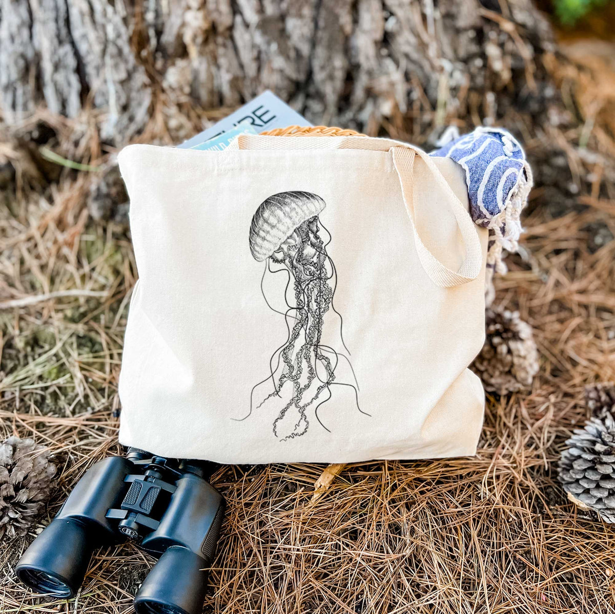 Canvas tote bag printed with a black ink illustration of a black sea nettle jellyfish with long trailing tentacles, resting on dry grass beside a pair of binoculars.