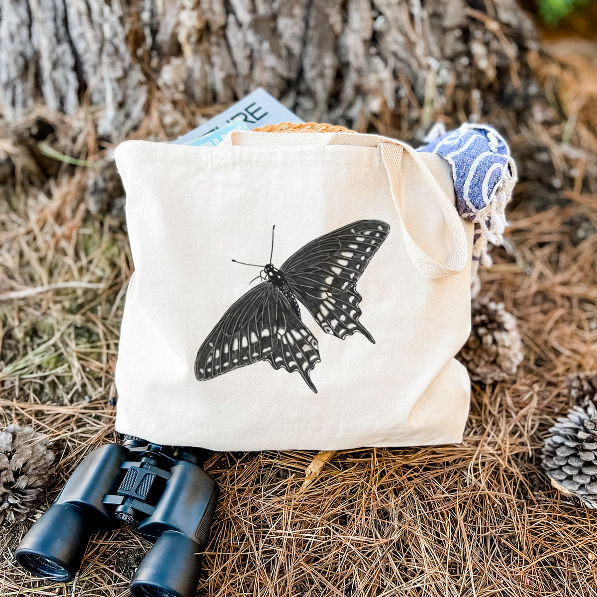 Canvas tote bag printed with a detailed black ink illustration of a black swallowtail butterfly with its wings spread, resting on dry grass beside a pair of binoculars.