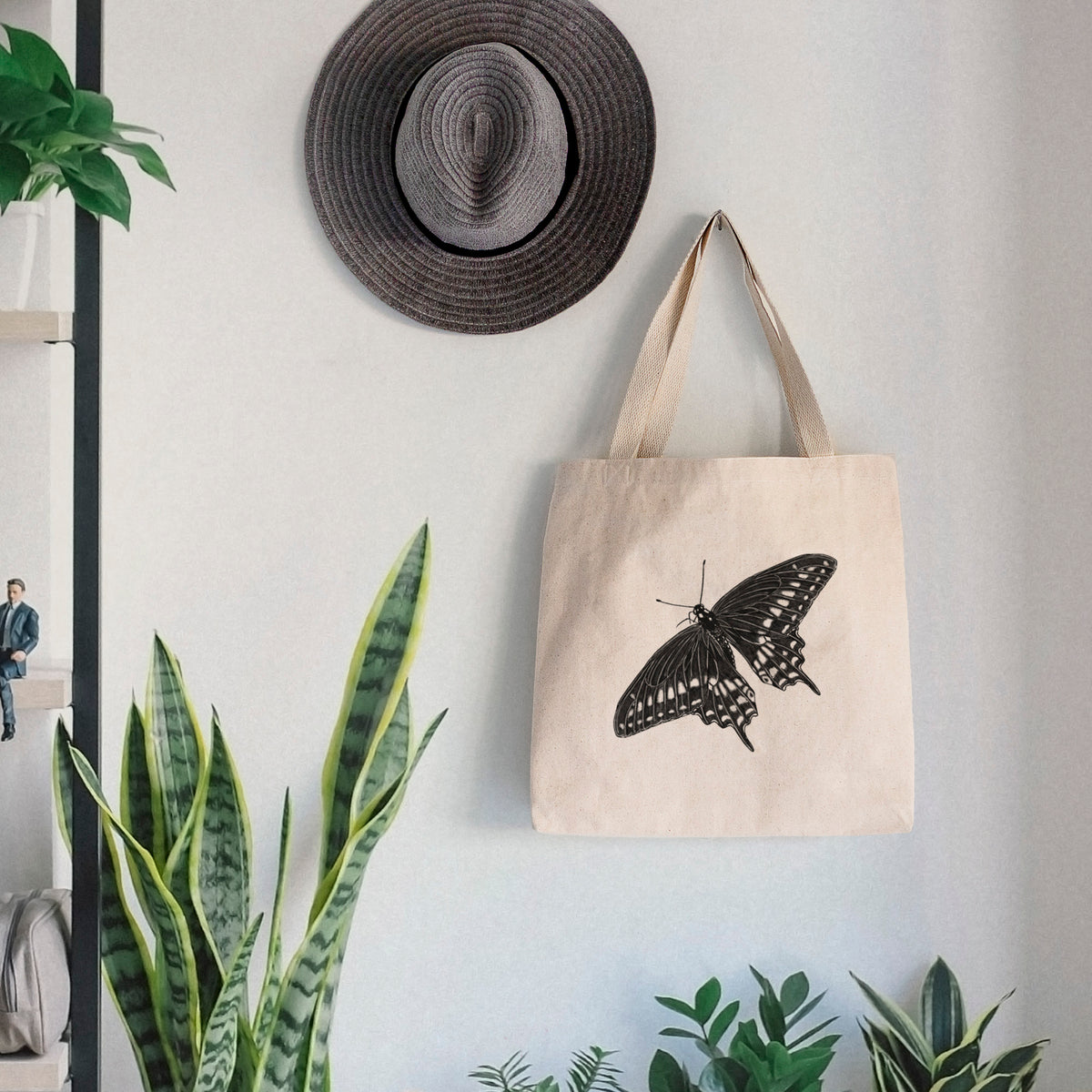 Canvas tote bag printed with a detailed black ink illustration of a black swallowtail butterfly with its wings spread, hanging on a wall hook below a hat, next to potted plants.